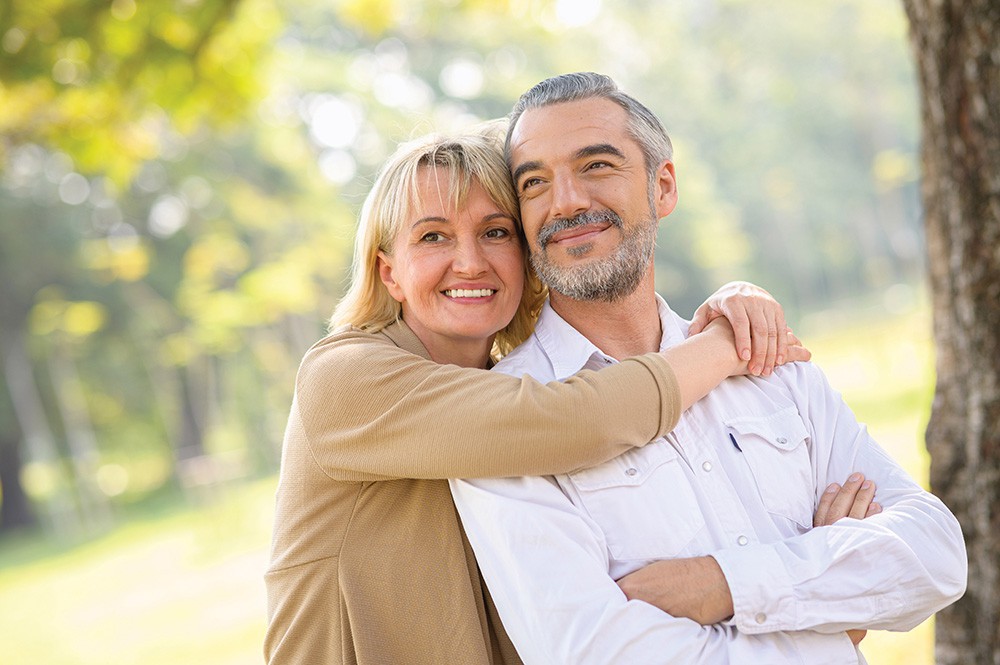 Women standing behind a man with her arms wrapped around his shoulders. They're outside and are older than 50